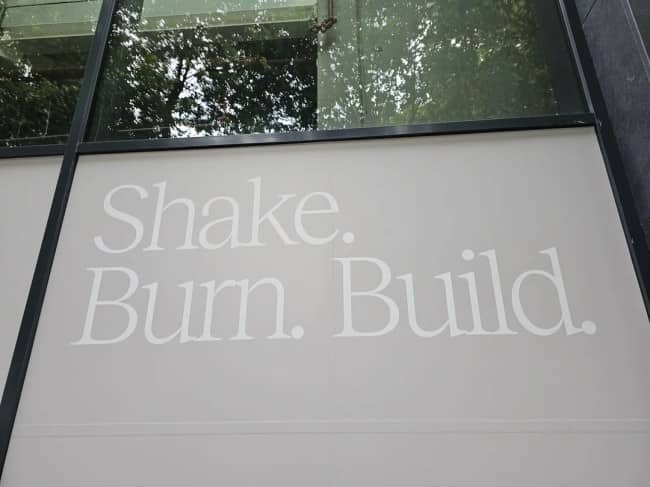 Glass window sign with white typography reading Shake Burn Build in a confusing font layout.