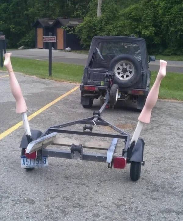 Empty boat trailer parked behind a vehicle featuring two mannequin legs attached upright on the sides.