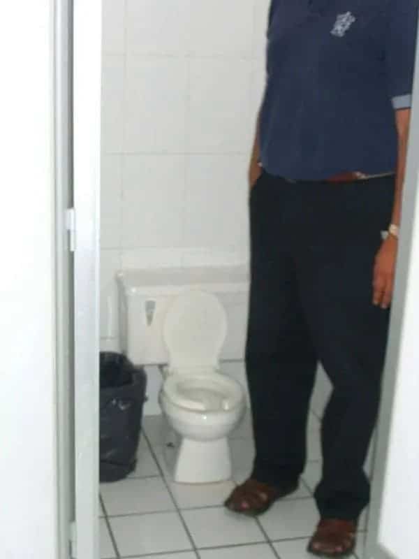 A tiny toilet installed inside a public bathroom stall next to a full-grown tall adult.