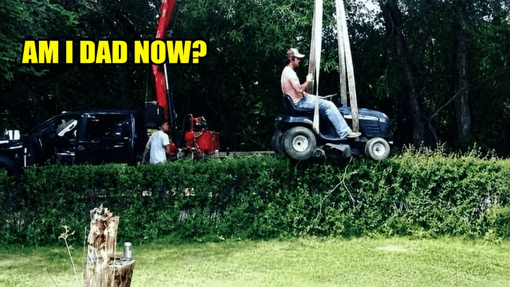 Man sitting on lawn mower suspended in midair by crane over tall green hedge.