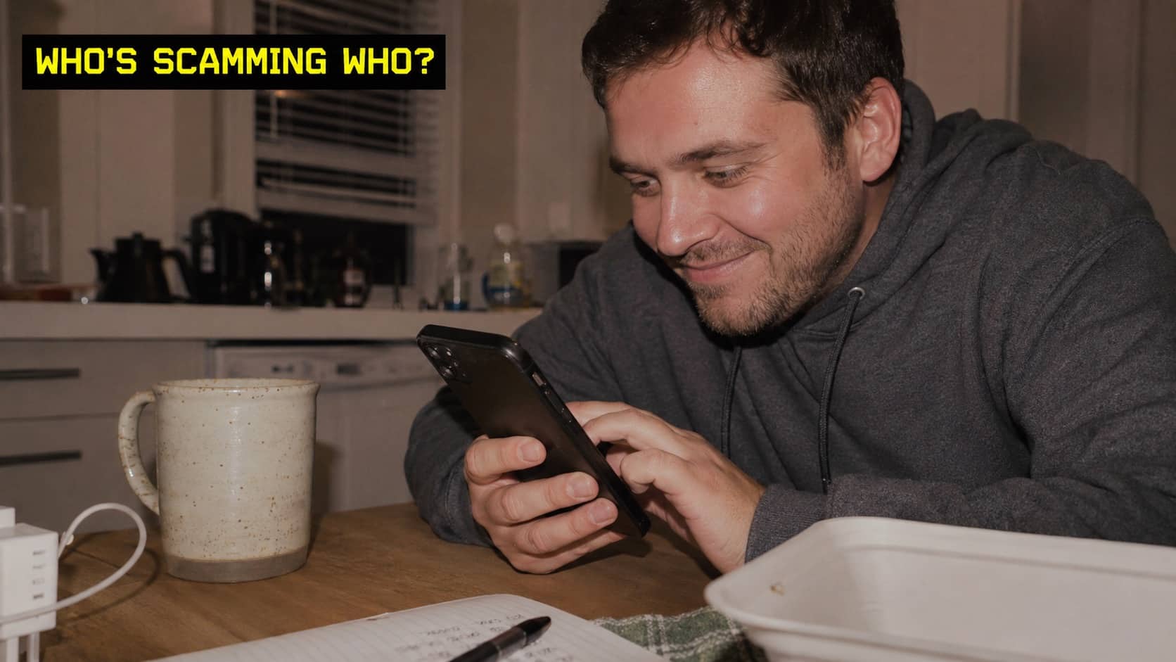 Smiling man sitting at kitchen table looking intently at his smartphone screen while holding pen.