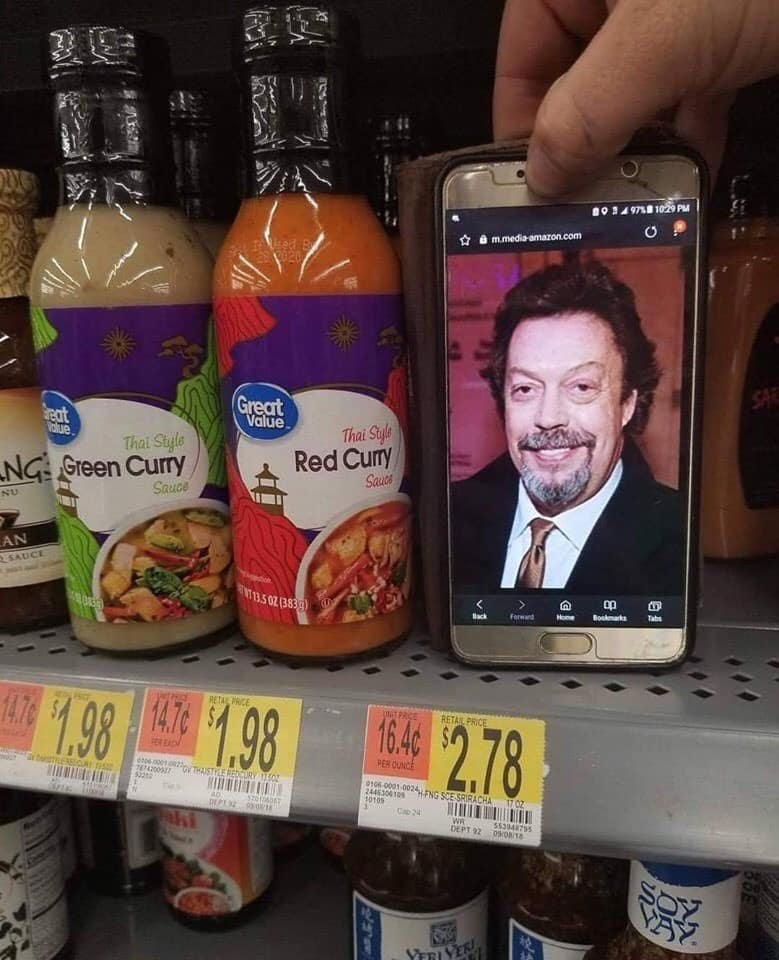 Grocery store aisle photo where a shopper holds up a smartphone displaying Tim Curry's face directly beside jars of Great Value Red Curry and Green Curry sauce.