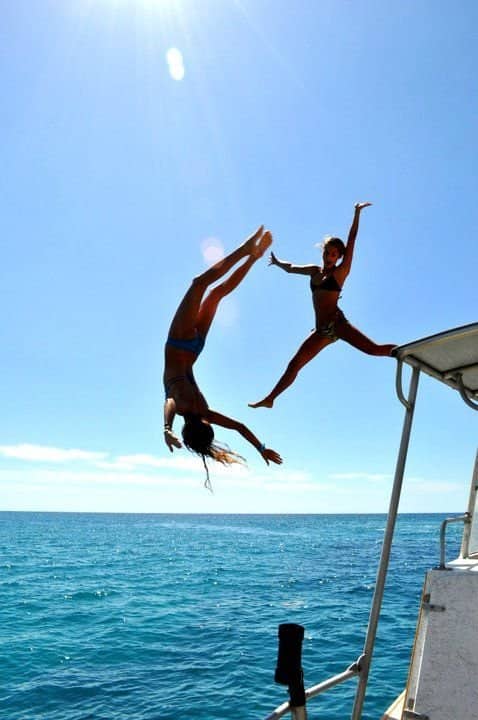 Two women diving headfirst off high boat roof canopy into ocean water.