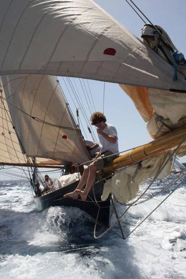 Young sailor sitting precariously on wooden bowsprit of high speed sailboat.