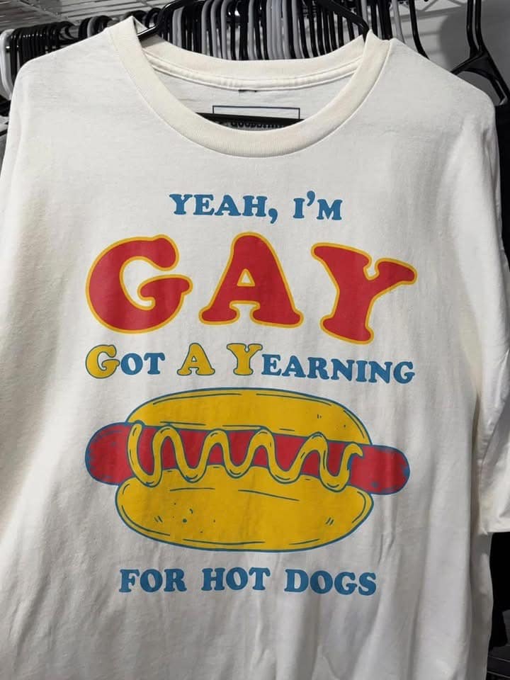 A white graphic T-shirt hanging on a rack with red and blue retro typography reading "YEAH, I'M GAY: Got A Yearning For Hot Dogs" above a mustard-covered frankfurter.