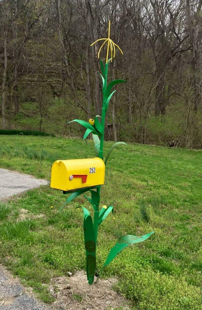 Bright yellow custom mailbox built into a tall green metal corn stalk in a grassy field.