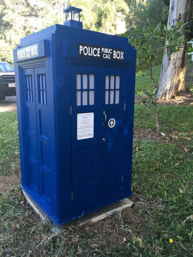Life-sized blue TARDIS police box custom mailbox sitting in a lush green front lawn space.