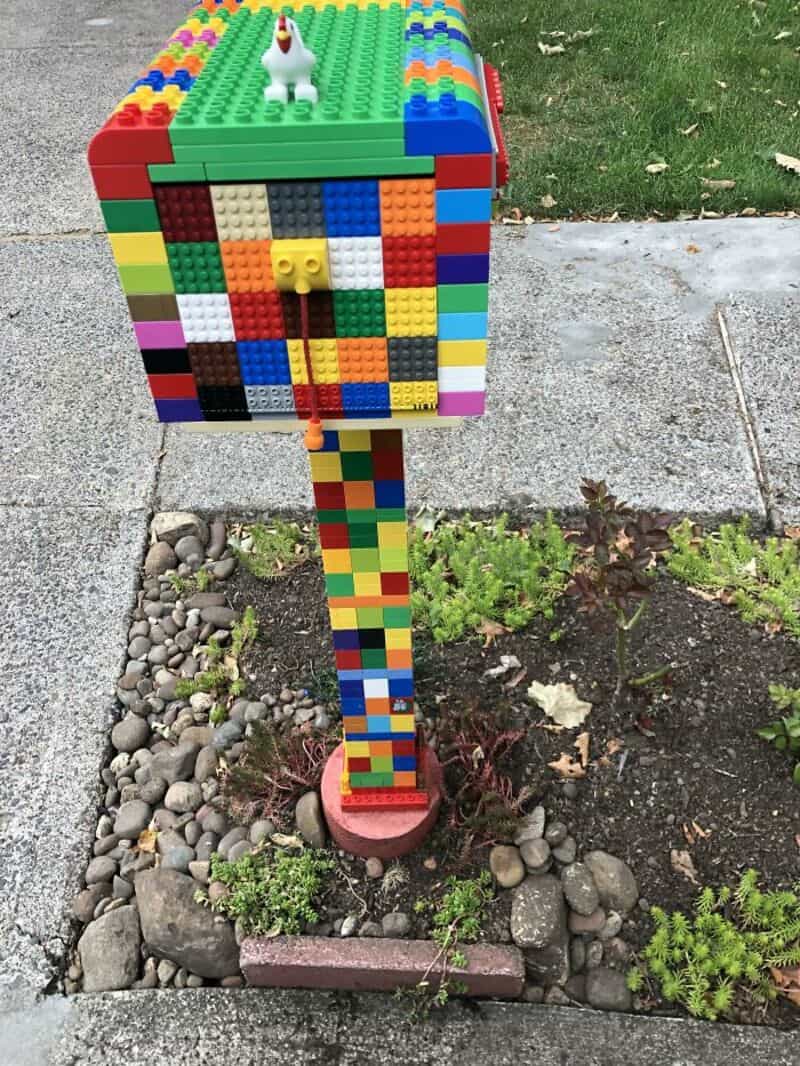 Colorful custom LEGO brick built mailbox standing in a rock garden beside a suburban sidewalk.