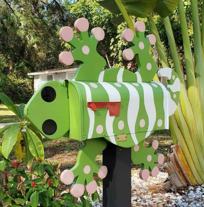 Large green spotted gecko lizard shaped cool mailbox attached to a post among tropical plants.