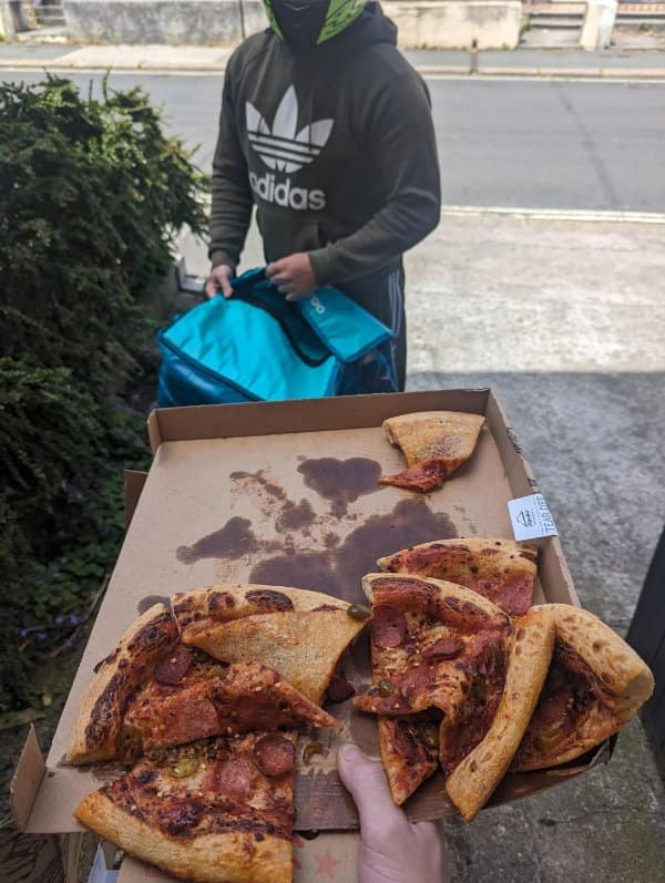 Cursed pizza delivery presentation showing an open box where the entire pie has violently slid into a jumbled, ruined heap of cheese and pepperoni along one edge.