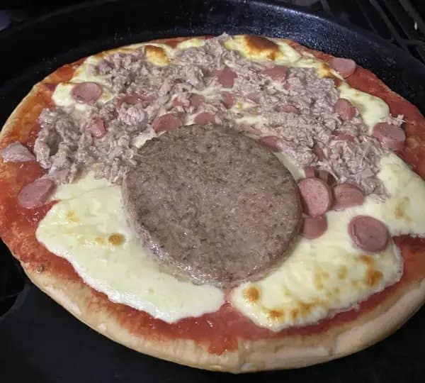 Cursed pizza cooked in a cast-iron pan, topped with canned shredded tuna, sliced hot dogs, and a whole precooked hamburger patty placed right in the center.