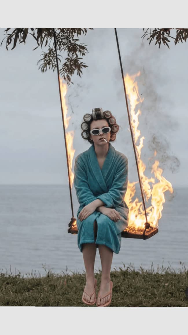 Woman in a bathrobe sitting calmly on a burning swing looking cute but terrifying.