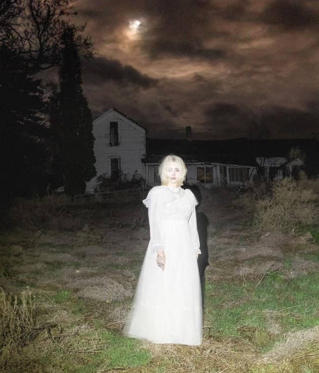 Ghostly blonde woman standing outside an abandoned house at night looking cute but terrifying.