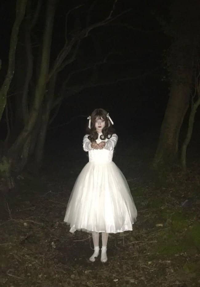 Girl in a white dress reaching hands forward in dark woods looking cute but terrifying.