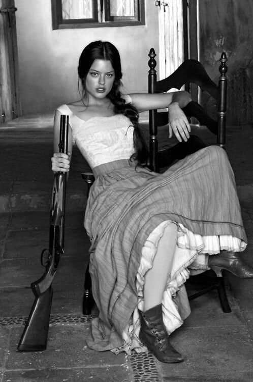 Vintage style woman in a long skirt casually holding a shotgun looking cute but terrifying.