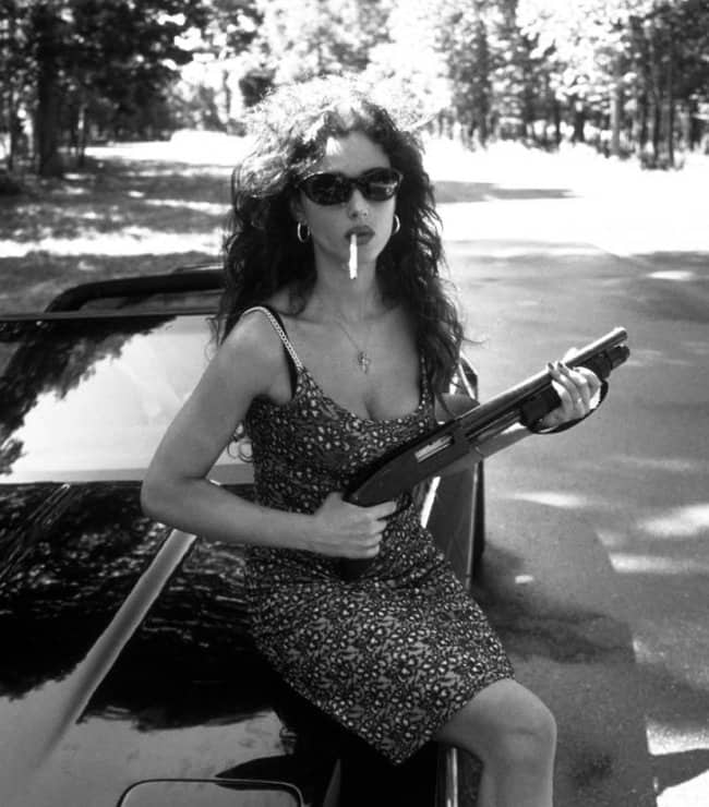 Woman in sunglasses smoking and holding a shotgun against a car looking cute but terrifying.