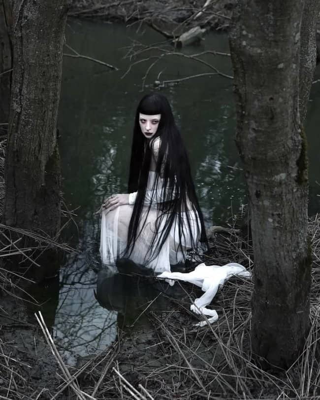 Pale goth woman with long black hair crouching in swamp water looking cute but terrifying.
