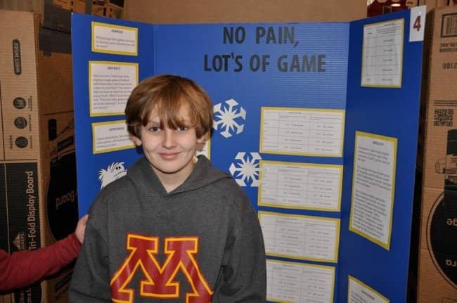Smirking boy standing in front of a blue science fair board titled No Pain, Lot's of Game.