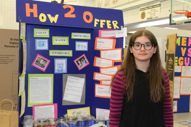 Girl standing beside a blue science fair presentation board titled How 2 Offer.