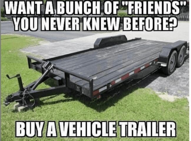 Large black dual-axle flatbed utility trailer parked on green grass lawn meme background.