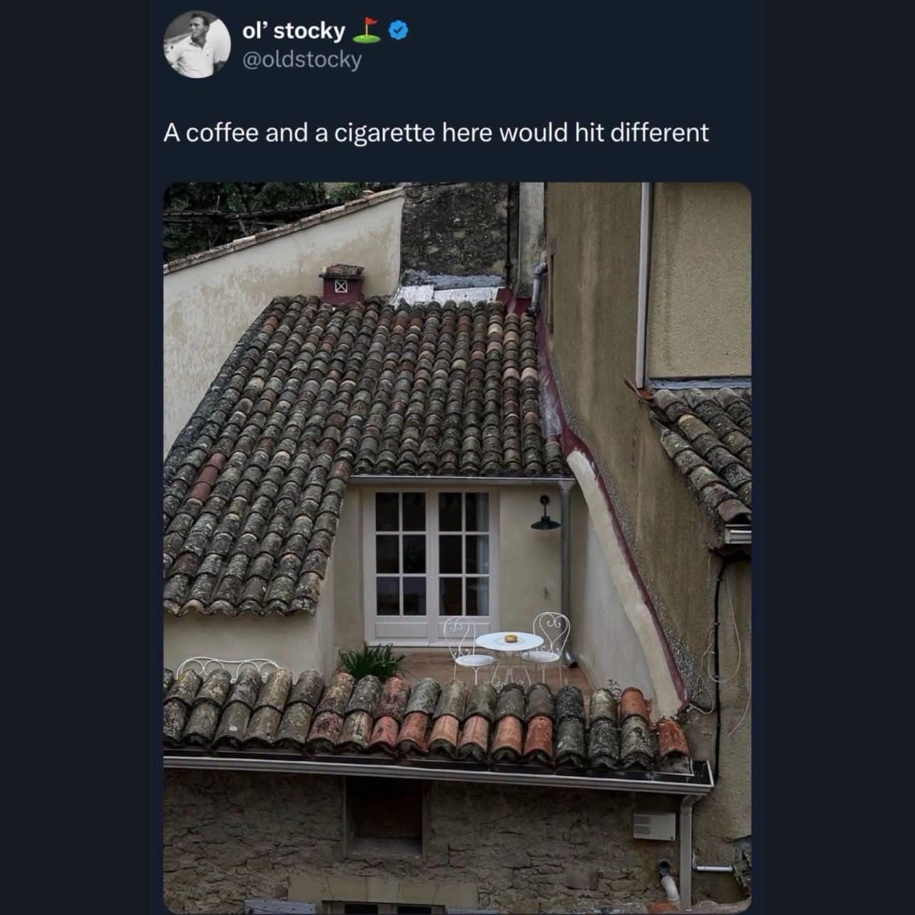 A cozy, secluded rooftop balcony with a small white table and two chairs wedged between European terracotta tile roofs, featured in a relatable meme dump tweet about drinking coffee.