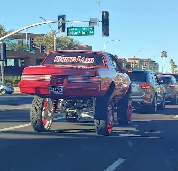 An over-the-top modified car photo acting as a hilarious meme showing a lifted red classic sedan on giant monster-truck suspension driving in traffic with a "YOUNG LAID" decal.