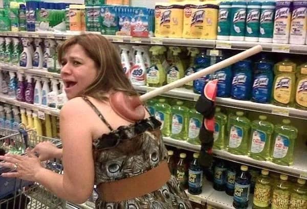 A woman in a grocery store cleaning supplies aisle turning around with a rubber toilet plunger suction-stuck firmly to her upper back.