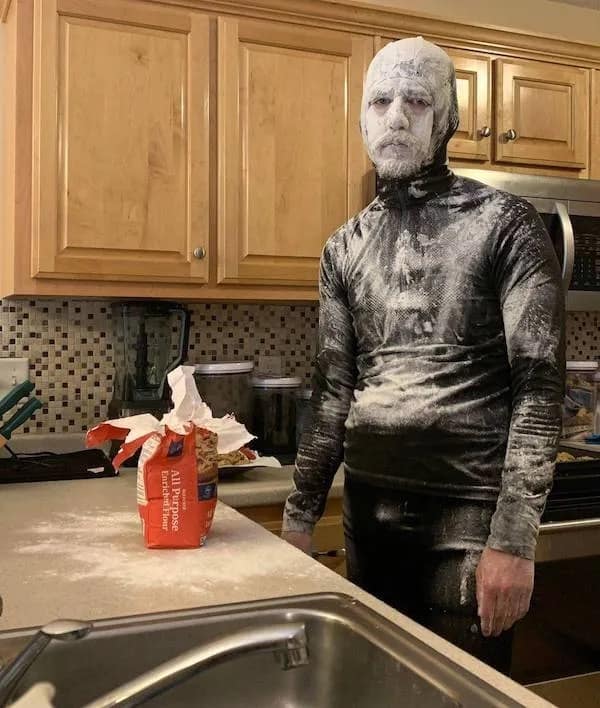 Covered head-to-toe in a thick layer of white baking flour, a man stands completely frozen in a kitchen next to an open flour bag, looking like a statue in a chaotic meme dump shot.