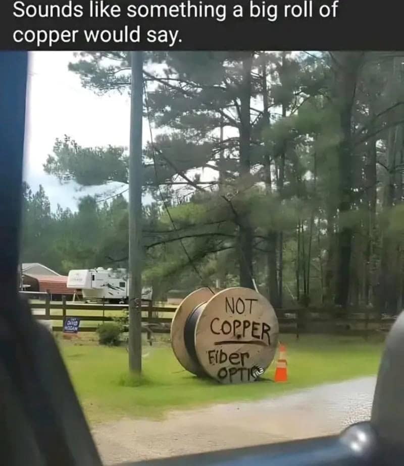 A large wooden cable reel parked on a rural lawn with "NOT COPPER Fiber Optic" hand-painted on it, captioned suspiciously in a meme dump.
