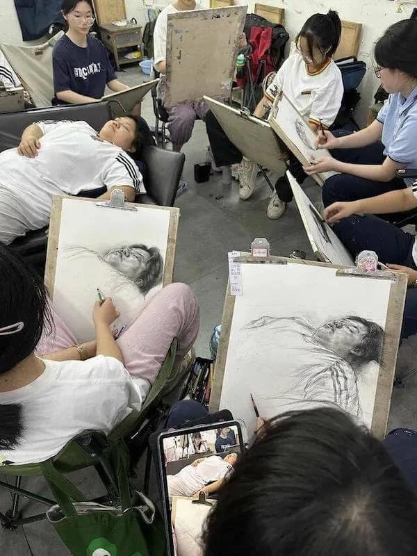 High-angle photograph of an art class where every student is drawing detailed charcoal portraits of a classmate sleeping on a lounge chair on the floor.