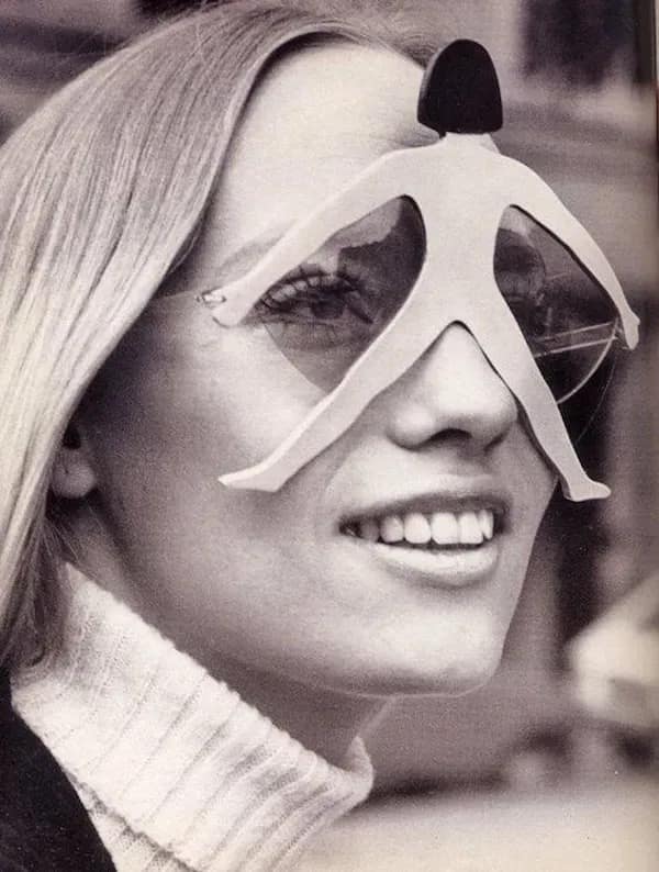 Vintage black-and-white fashion portrait of a smiling woman wearing eccentric white sunglasses styled in the shape of a spread-eagled stick figure spanning across her forehead and eyes.