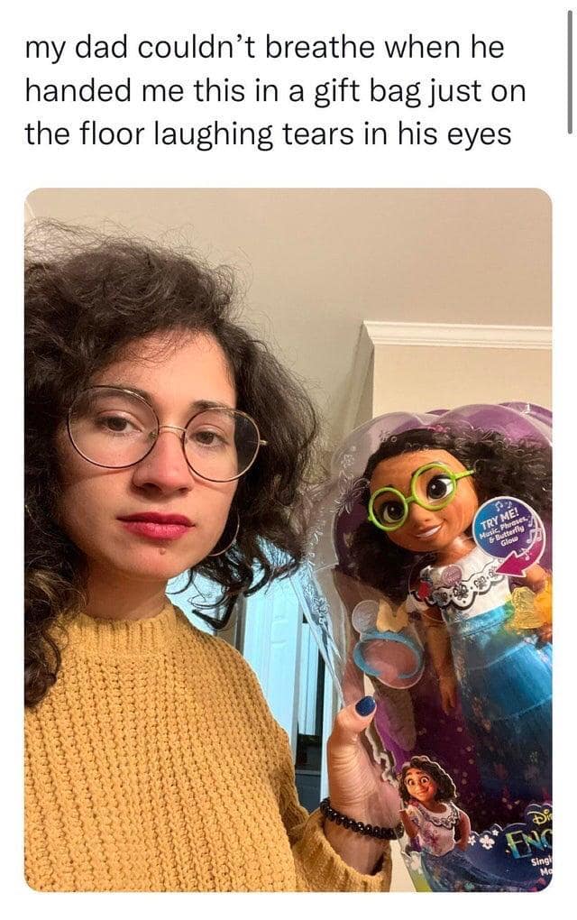 A woman with curly hair and round glasses holding a boxed Disney Encanto Mirabel doll gifted by her laughing father.