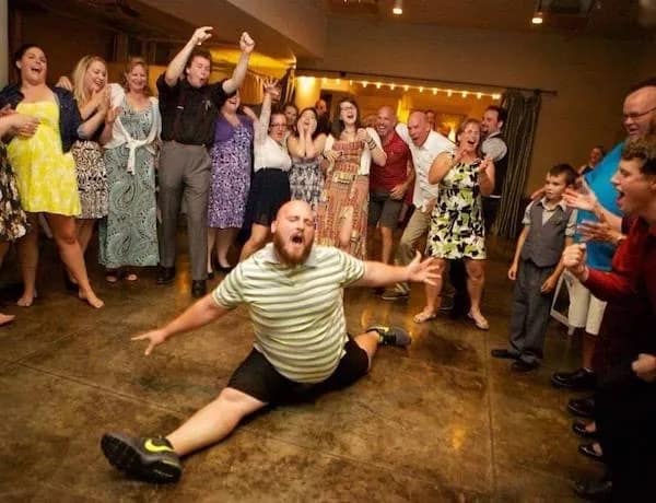 A bearded man performing a full gymnastic split in the middle of a wedding reception dance floor surrounded by cheering guests.