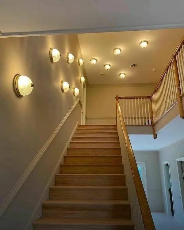 A chaotic interior design nightmare featured as a hilarious meme showing a staircase area illuminated by dozens of randomly placed wall sconces and ceiling dome lights.