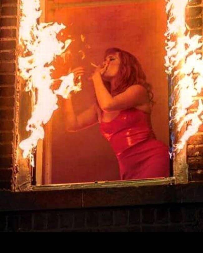 Woman in red dress lighting cigarette off burning window frame inside flaming building.