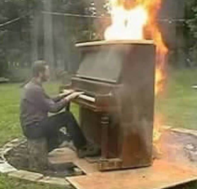 Man playing upright piano that is fully engulfed in flames in backyard.