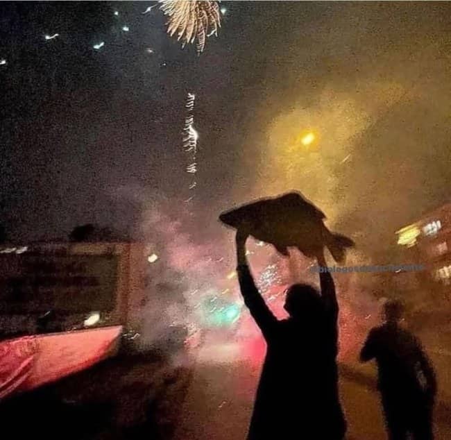 Silhouetted person holding giant dead carp high overhead during explosive nighttime fireworks.