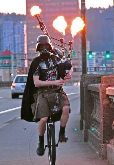 Person in Darth Vader costume riding unicycle while playing flaming bagpipes outdoors.