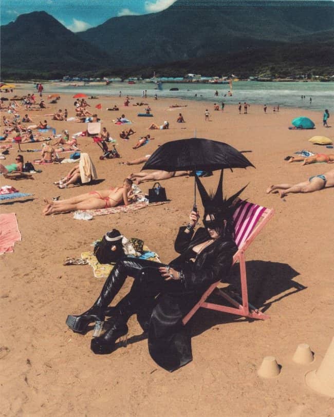Goth person dressed in full black leather sitting under umbrella on crowded sunny beach.
