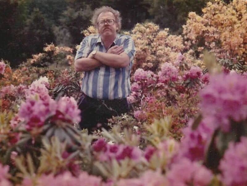 Old school cool man with glasses standing discontentedly surrounded by colorful blooming flowers.