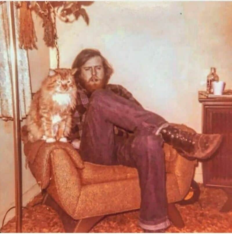 Old school cool man sitting in an armchair next to a fluffy orange cat.
