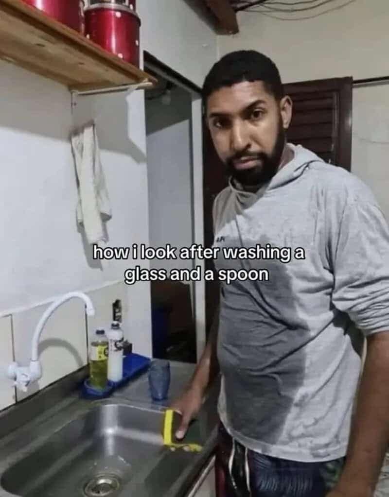 A man standing in front of a kitchen sink with his grey hoodie completely soaked across the chest after washing a single glass and spoon.