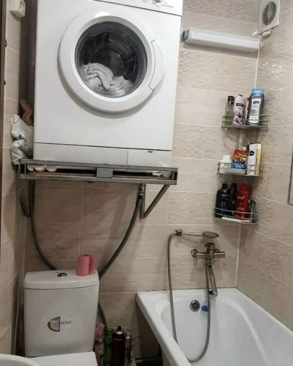A heavy washing machine resting on a wall shelf installed directly above a toilet.