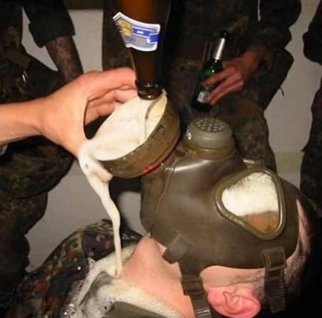 Soldier wearing a gas mask while another soldier pours beer straight into it.