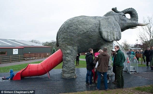 Families standing near a red slide protruding from an elephant rear at the worst playgrounds.
