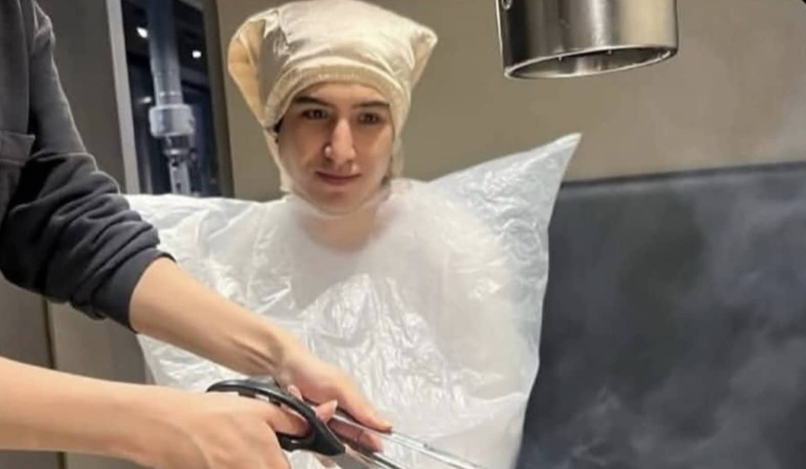 Person wearing a plastic protective suit and hairnet sitting under salon equipment for styling.