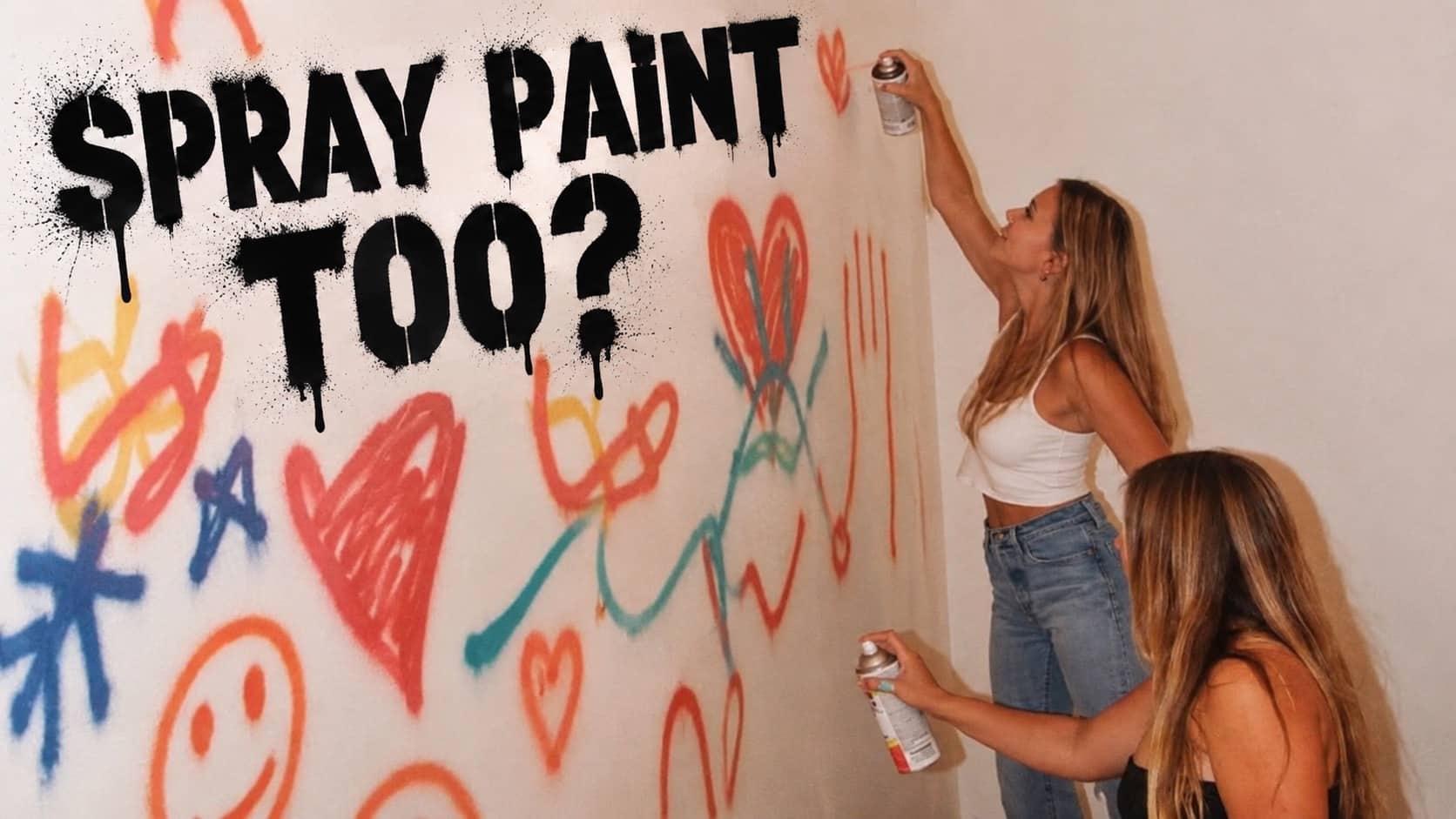 Two women painting colourful graffiti shapes and heart artwork on a white indoor wall.