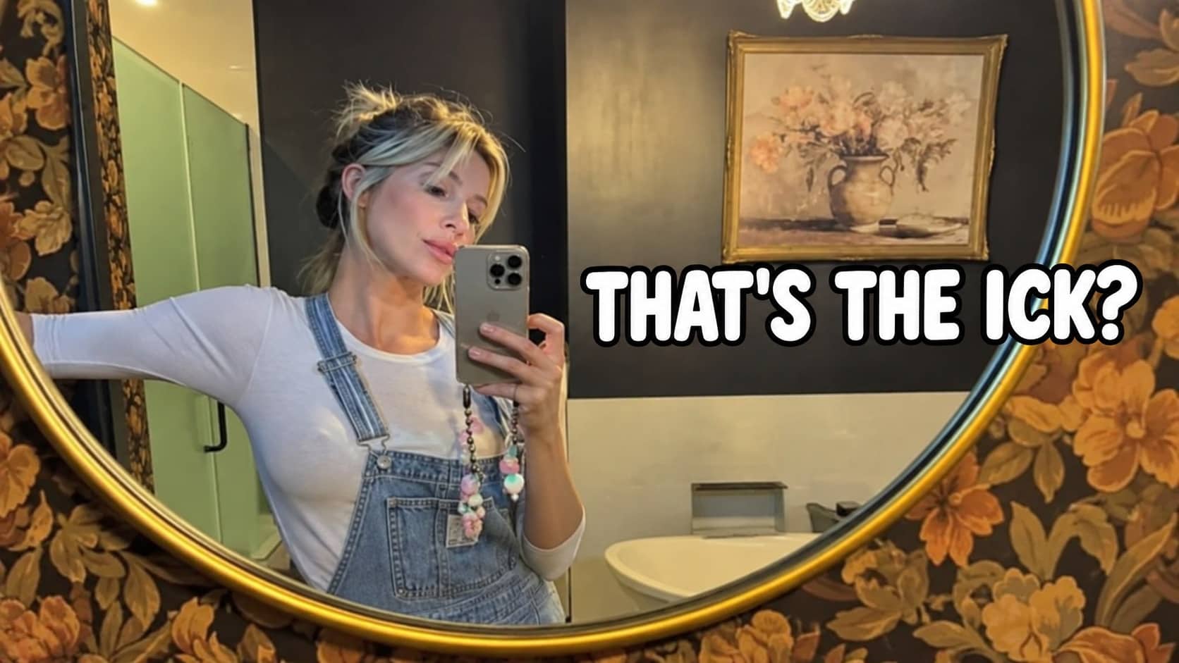 Woman in denim overalls taking a mirror selfie in a floral vintage styled bathroom.