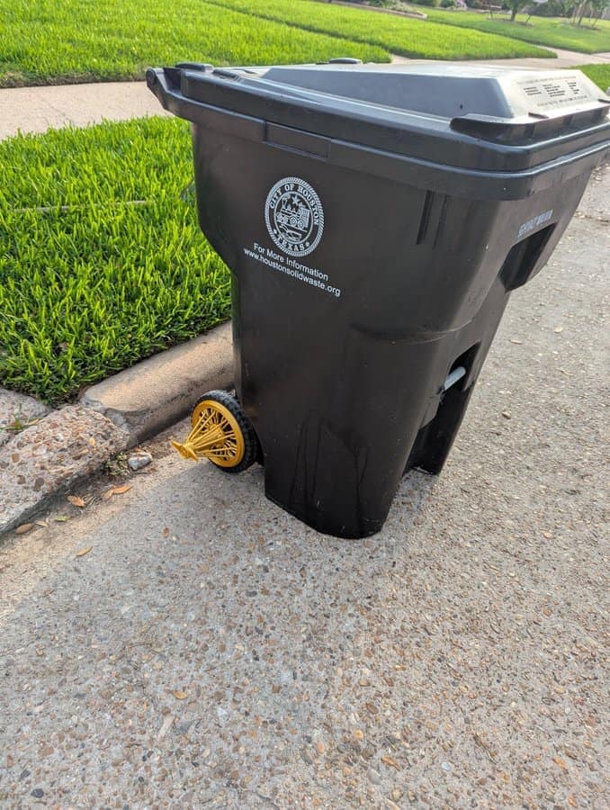 Garbage bin on the curb fitted with a shiny gold spinner rim wheel.