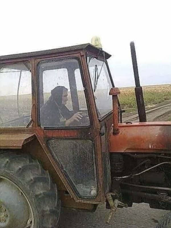 Elderly Balkan babushka in black headscarf driving red agricultural tractor through rural farm field.